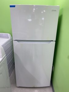 Frigidaire white two-door refrigerator with top freezer, taped labels, in a bright green room.