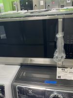 Showroom scene with a large black LG washer/dryer unit in the foreground, wrapped in plastic, surrounded by other appliances in a showroom with green walls. A person in white stands in the background.