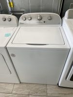 Row of white laundry machines in a laundromat; center washer has a Whirlpool control panel, flanked by other machines and a metal wall behind.