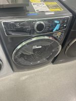 Front-loading Electrolux washing machine in a showroom, covered in plastic wrap with blue tape and energy-guide sheets on top of the unit.