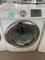 Front-loading Samsung washing machine with a circular glass door and a control panel; green sticky note attached to the left side of the console in a showroom.
