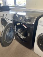 Front-loading washing machine with its door open in a showroom, adjacent dryer visible behind it.