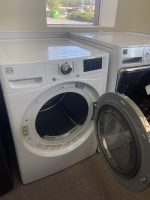 Front-loading washing machine with the door open, shown in a bright showroom alongside a dryer.