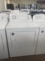 Row of white Amana top-load washing machines in a showroom, with sale signs on the lids and a brown carpet floor.
