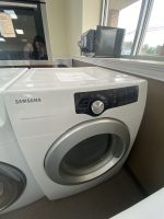 White Samsung front-load washing machine in a laundromat with a microwave on the windowsill and a person reflected in the glass door of the washer.