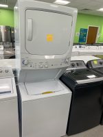 White front-loading dryer stacked on a matching washer in a showroom, with a yellow sticky note on the dryer door and bright green walls in the background.