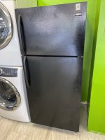 Black textured two-door refrigerator (top freezer, bottom fridge) with Frigidaire badge, shown next to a silver washing machine against a bright green wall in a showroom.