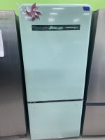 Mint green retro refrigerator with a chrome nameplate and a colorful metal flower decoration on the top left, shown between other appliances in a showroom.