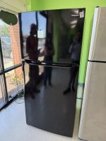 Black Frigidaire refrigerator on display in a showroom with a lime-green wall and a sticky note on the door; reflections of people in the glossy surface.