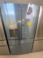 Two blue stainless steel side-by-side refrigerators in a showroom; left unit has an ice dispenser, right has a flyer taped to the door.