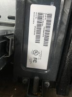 Label on an industrial device showing barcodes, model and serial numbers, mounted on a black metal housing with screws nearby