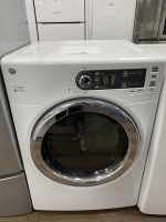 White GE front-loading washing machine with a chrome circular door in a showroom, control panel on the right and price-tagged appliances in the background