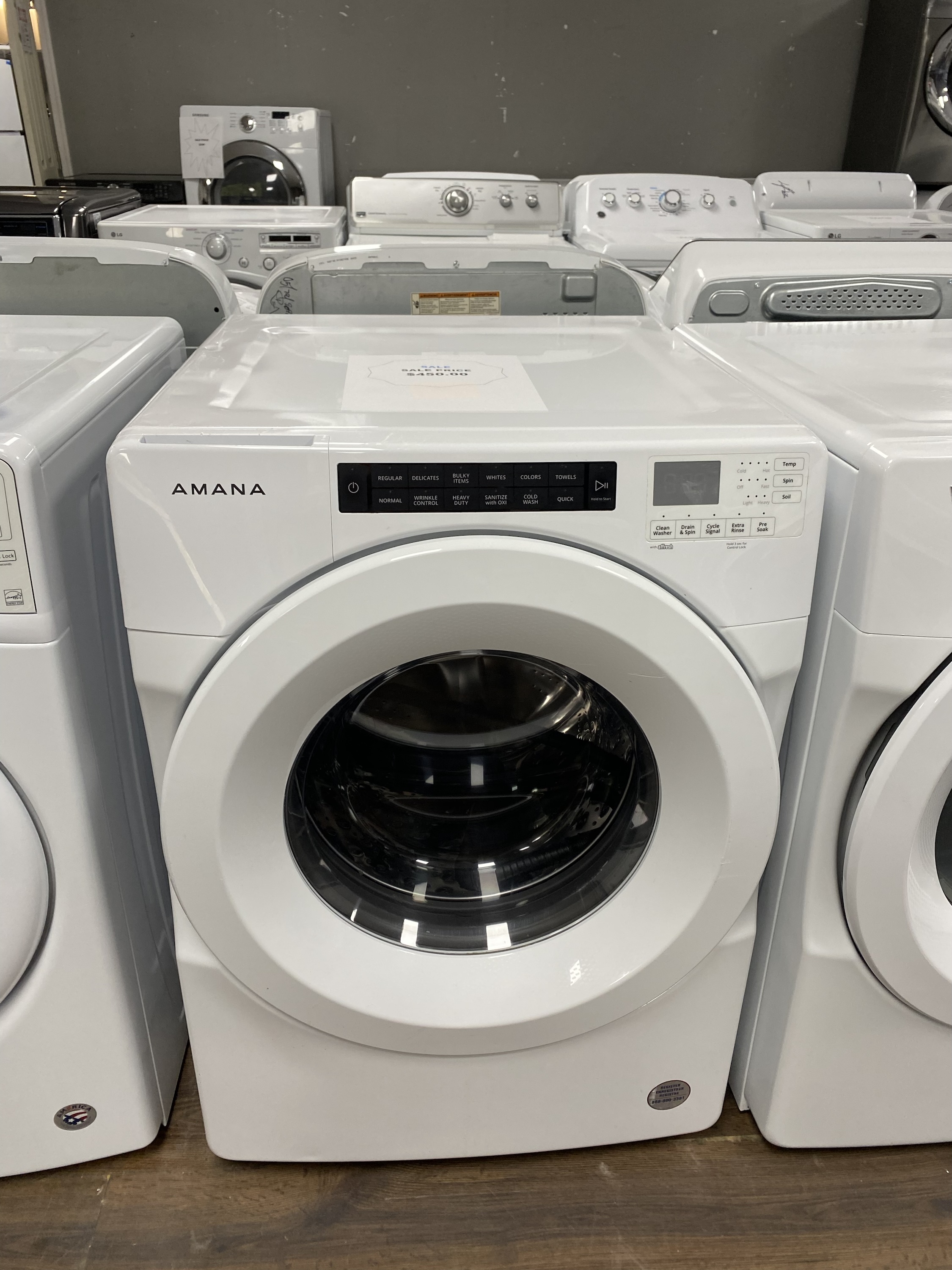 White Amana front-loading washing machine on display in a showroom with other laundry appliances in the background.”,