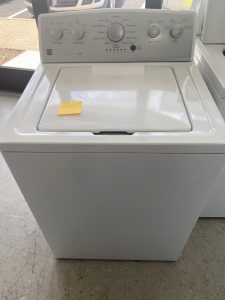 Top-loading washing machine with closed lid and a sticky note on the lid reading 'colonial' in a laundromat setting.