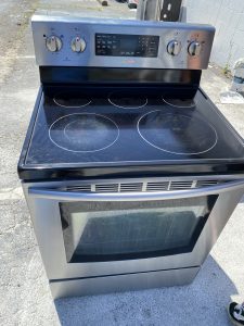 Stainless steel electric range with four-burner glass cooktop and digital control panel outdoors, oven door visible below.