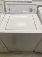 White top-loading washing machine with its lid closed and a circular cycle dial on the control panel in a laundry room setting