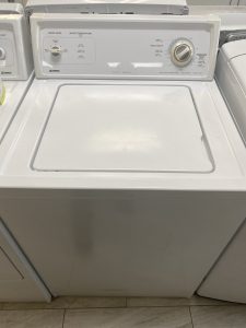 White top-loading washing machine with its lid closed and a circular cycle dial on the control panel in a laundry room setting