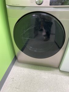 Front-loading washing machine with a cracked glass door against a lime-green wall. Control panel at the top is visible.