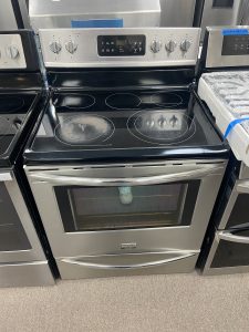 Stainless steel electric range with a black glass cooktop and six burner zones in a showroom setting