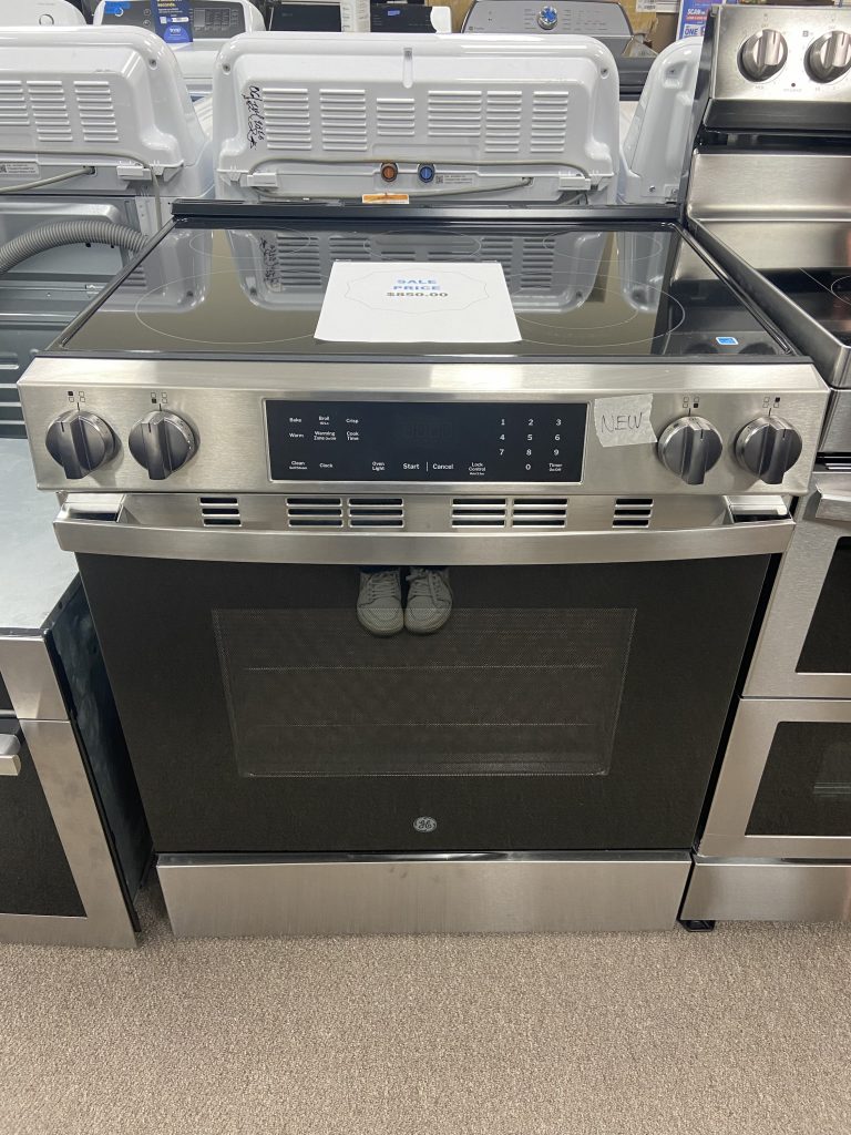Stainless steel GE electric range with a glass-ceramic cooktop on display; a price sheet sits on the cooktop.
