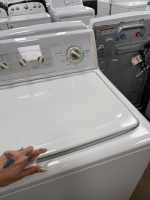 White top-loading washing machine in a showroom, a hand resting on the lid.